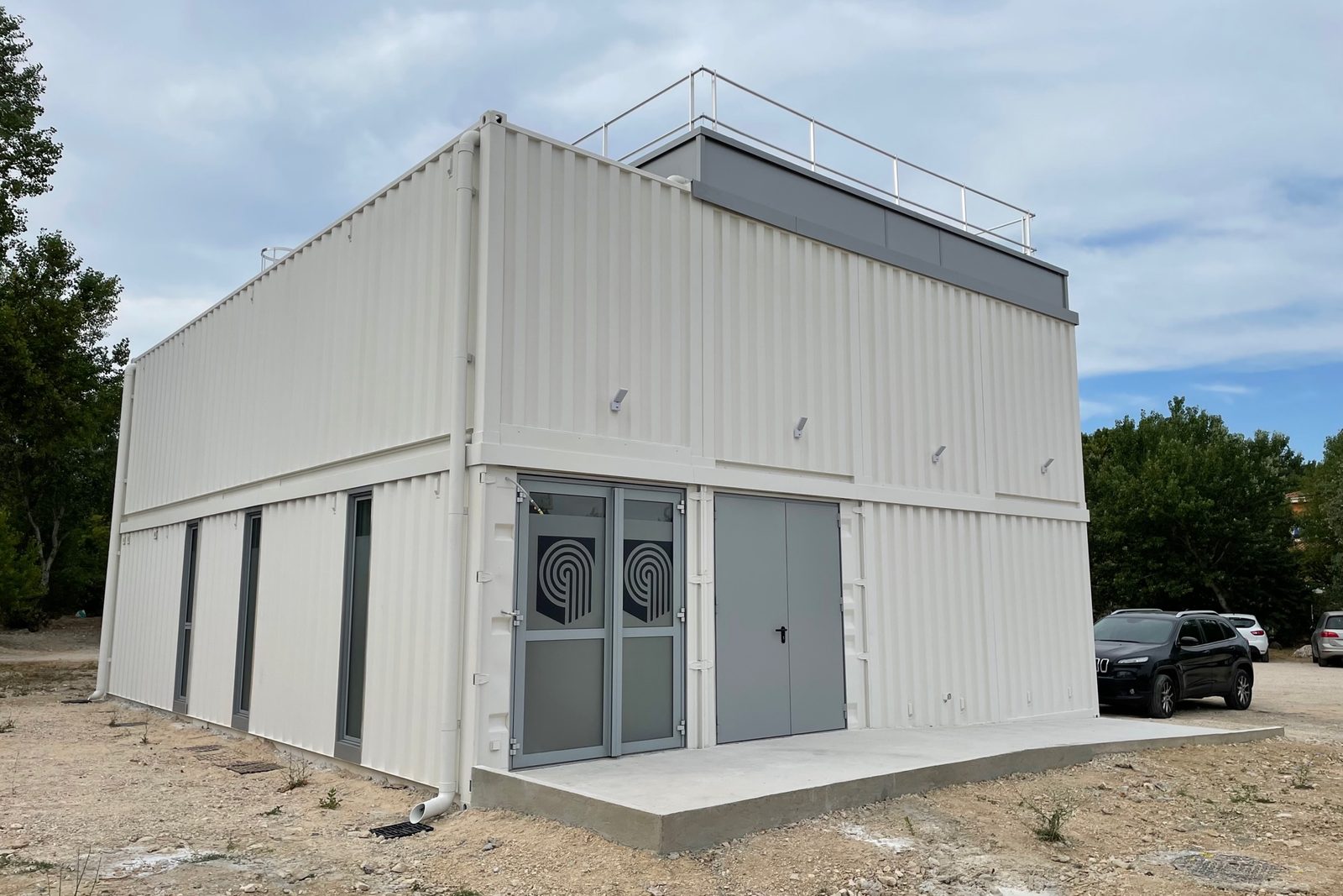 Salle de répétition et de spectacle en containers blancs empilés sur deux niveaux, avec double porte d'accès et rampe béton