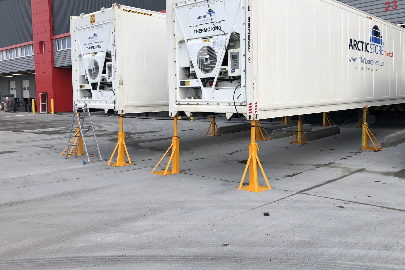 Containers frigorifiques 40 pieds posés sur des chandelles métalliques jaunes, devant un entrepôt